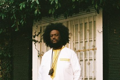 Kamasi Washington, a Black man dressed in a white shirt with a beard and voluminous hair, is pictured standing outside in front of a window. Green vines hang above the window.
