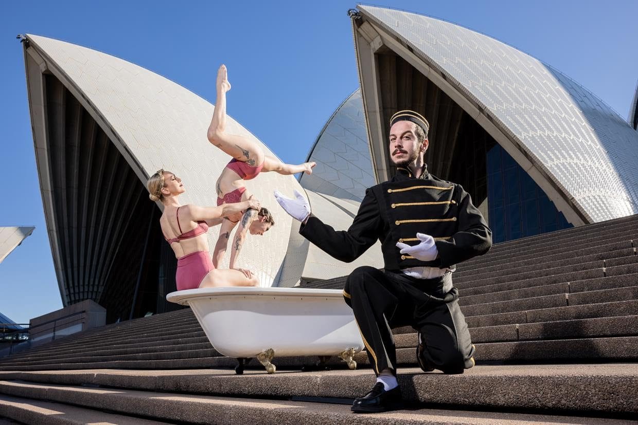 Two women in lingerie, CJ Shuttleworth and Bri Emrich, hold acrobatic poses in a bathtub on the Opera House's Monumental Steps with the Sails looming in the background. A man dressed as a hotel porter, Brendan Maclean, gestures to them from the foreground of the image.