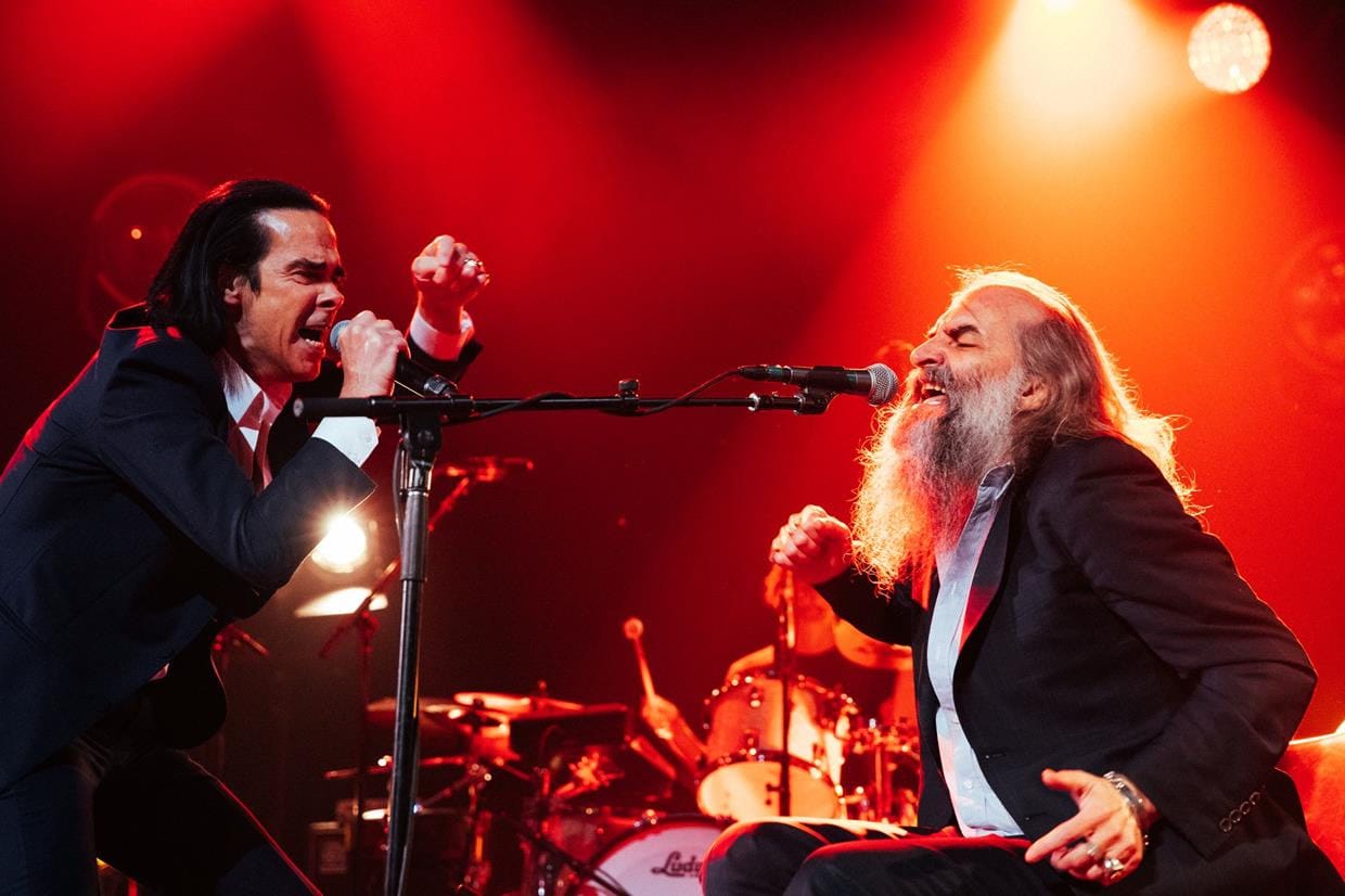 Nick Cave and Warren Ellis, two white men in black suits on stage under red lights passionately sing and gesture with outstretched arms and clenched fists.The atmosphere is bold and dramatic. Image credit: Megan Cullen