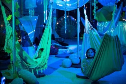 Two children of late primary school age are pictured relaxing in hammocks with headphones on during the creative development of After The Flood in the Centre for Creativity. The image is bathed in blue light while other reflective items hang from the ceiling beihind them in the installation. PHOTO CREDIT: Cassandra Hannagan