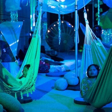Two children of late primary school age are pictured relaxing in hammocks with headphones on during the creative development of After The Flood in the Centre for Creativity. The image is bathed in blue light while other reflective items hang from the ceiling beihind them in the installation. PHOTO CREDIT: Cassandra Hannagan