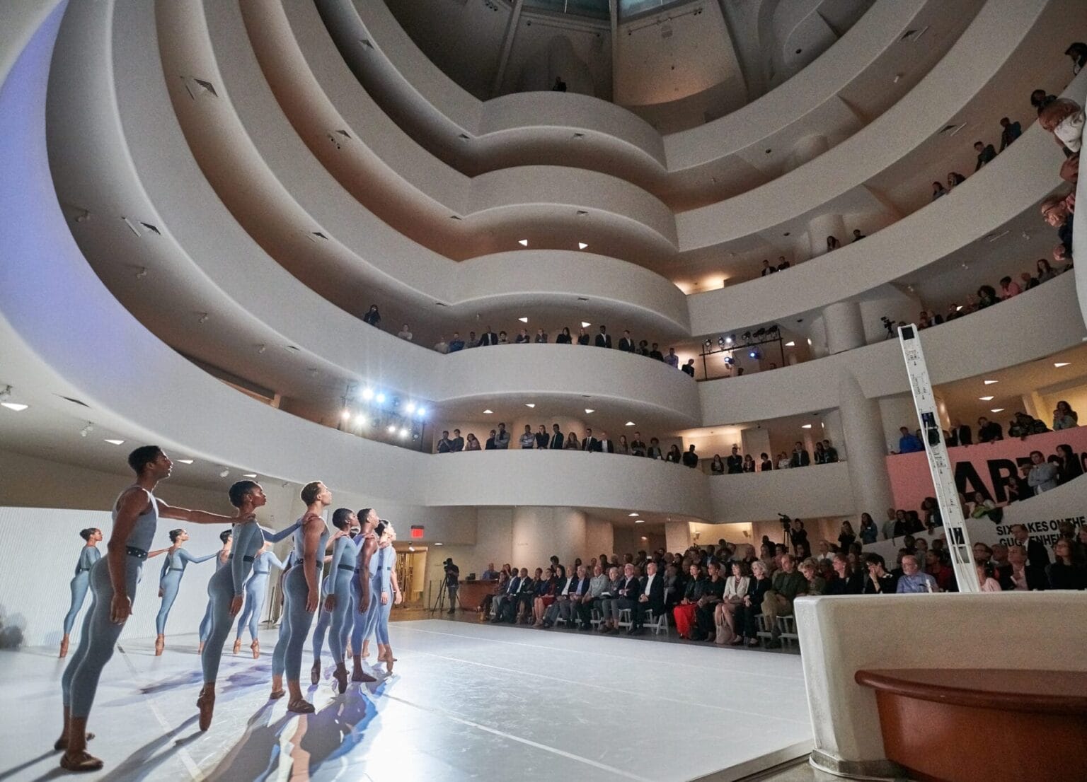 Performers in the middle of the Guggenheim, standing in a line with arms raised. Audience members stand and watch along the spiral staircase surrounding them