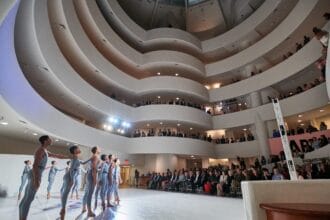 Performers in the middle of the Guggenheim, standing in a line with arms raised. Audience members stand and watch along the spiral staircase surrounding them