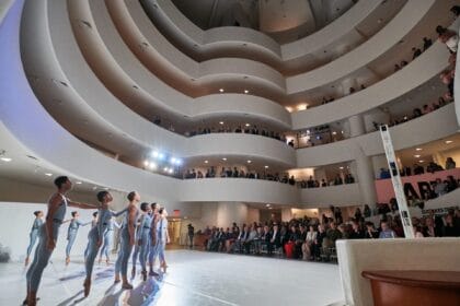 Performers in the middle of the Guggenheim, standing in a line with arms raised. Audience members stand and watch along the spiral staircase surrounding them